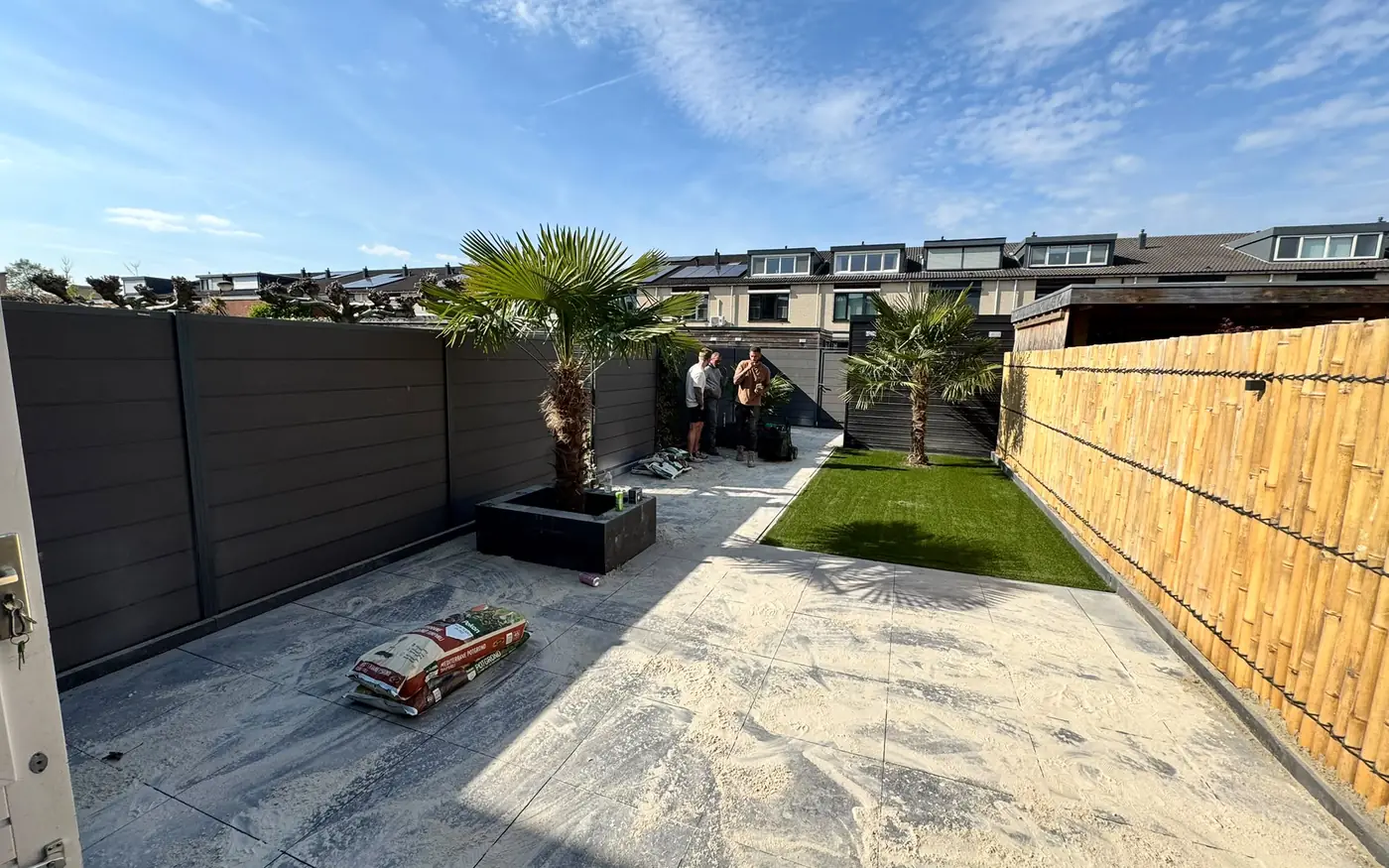 De afgeronde tuin met gazon, palmen en bestraat terras onder een blauwe lucht
